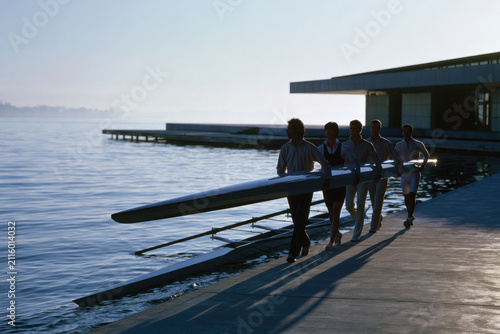 Five rowers carry their sleek boat to the dock in early morning mist, depicting the dedication and effort required in rowing sports before the sun rises.