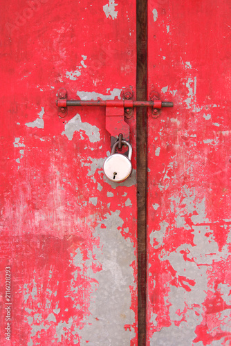 Close-up of a weathered red metal door secured with a padlock, featuring peeling paint and a rustic textured surface