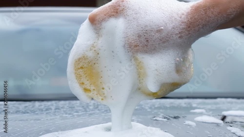 Close-up of a hand squeezing a soapy sponge over a car, releasing thick foam during vehicle washing, highlighting automotive detailing, cleaning, and maintenance.