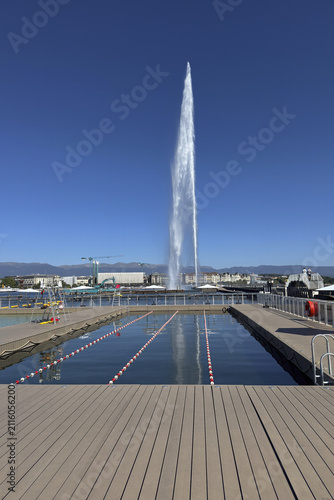 A tall jet of water rises over Lake Geneva, with people walking along the pier beneath the clear summer sky.