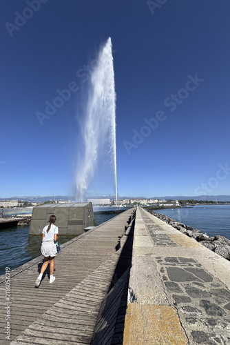 A tall jet of water rises over Lake Geneva, with people walking along the pier beneath the clear summer sky.