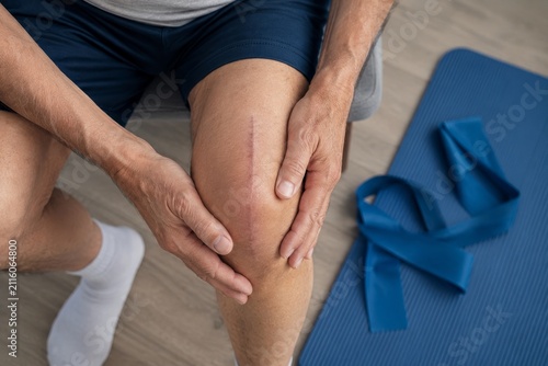 Senior man touching knee with surgical scar after joint replacement surgery. Patient doing physical therapy with resistance band for rehabilitation of knee injury. Orthopedic medical recovery.
