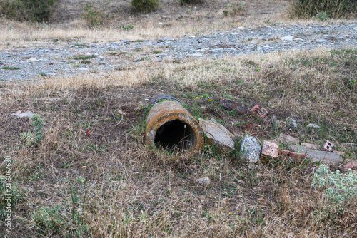Cuadro en lienzo a weathered, cylindrical drainage pipe protruding from a dry, grassy embankment next to a gravel path, with scattered stones and debris nearby