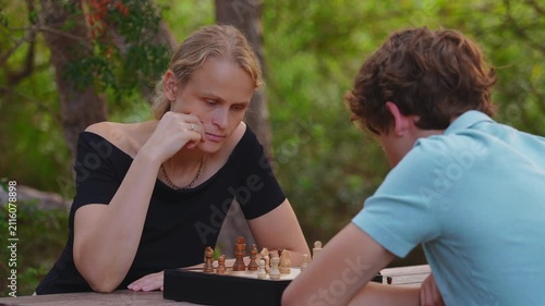 Mother and her fifteen year old teenage son play chess outdoors in a park, with the mother thinking deeply about her next move on the wooden board