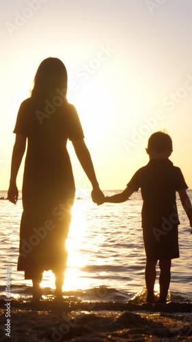 Mother and son walk toward the sea at sunset while holding hands. Warm silhouette captures family travel, shared adventures, unforgettable vacation memories and joyful moments together.