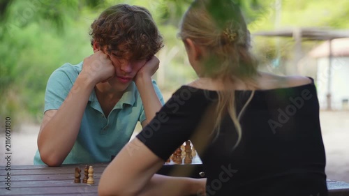 A teenage boy with curly hair concentrates on a chess game, sitting opposite his mother at a wooden table in an outdoor park