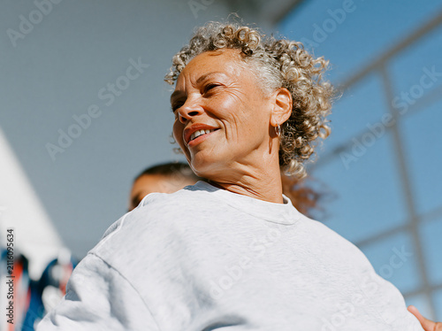 Happy senior woman with gray curly hair smiling in bright sunlight during a fitness class, joyful healthy aging and vitality concept.