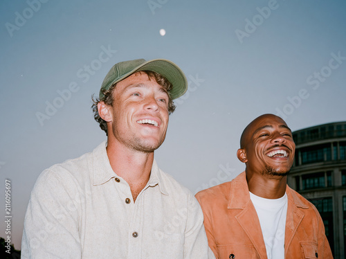 Two multiethnic male friends laughing together on a city rooftop at dusk, candid flash photo with authentic joy and urban skyline background.