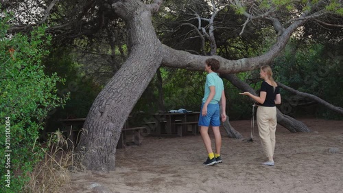 A mother and her teenage son spend quality time together in a pine forest park during a summer camping trip, talking and playing under a large low hanging tree branch