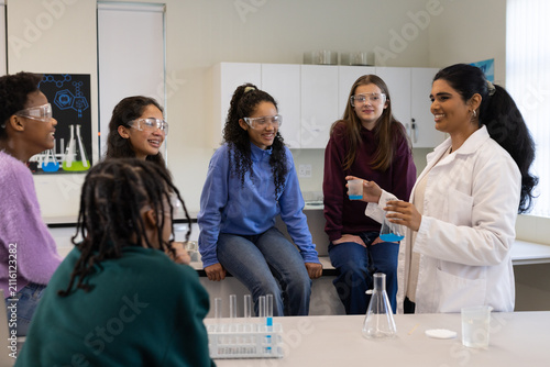 Indian teacher labcoat holding blue beakers, showing chem in lab to 5 goggle-clad teens, copy space