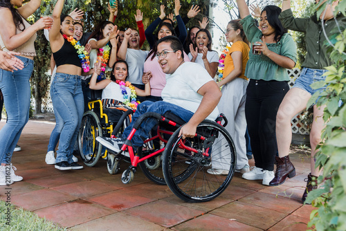 latin people in wheelchair with disabilities having fun and dancing surrounded of group of hispanic friends in party outdoors in terrace in Mexico Latin America in social inclusion concept