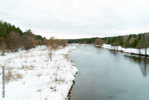 River flows through a snowy landscape with trees along the banks on a cloudy day