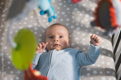 Baby lying in crib and looking up at hanging colorful mobile toys in a cozy nursery.