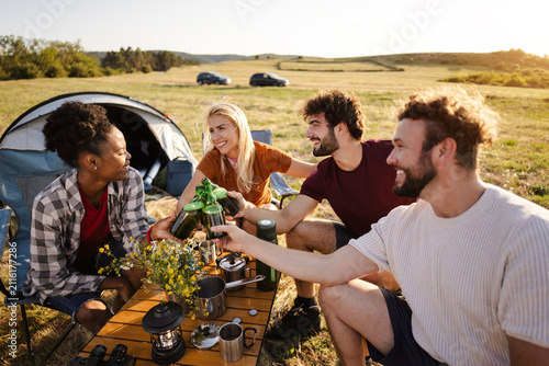 A group of friends enjoys drinks and camaraderie around a camping table in a vibrant meadow, basking in the golden hour light.