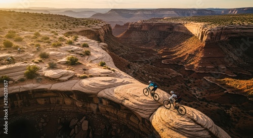 Two mountain bikers navigating a narrow rocky trail across a vast desert canyon at sunrise.