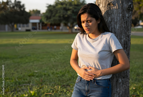 Young woman experiencing stomach pain and discomfort, leaning against a tree in a park setting