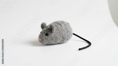 A small gray fuzzy mouse-shaped cat toy with a thin black tail, resting on a plain white surface, soft studio lighting highlighting the soft felt texture against a minimalist neutral background for