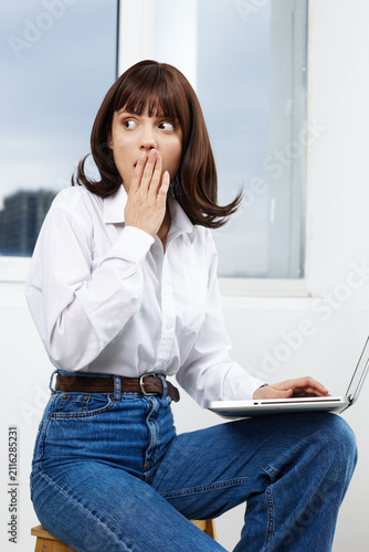 Fotografía A young woman sits indoors with a laptop, glancing aside with a surprised expression, suggesting a secret moment or unexpected discovery during work or study