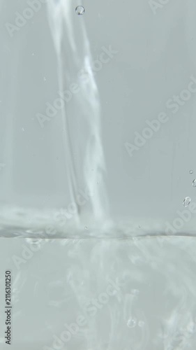 A stream of clear water pours into the container as the clear liquid spreads across the surface, mixing and swirling into a textured wavy pattern above the calm, clear water below.