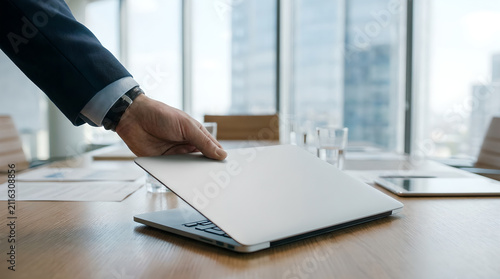 Businessman in a sharp suit closes his sleek silver laptop while sitting at a polished wooden conference table in a bright, modern office with expansive floor-to-ceiling glass windows.