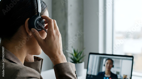Professional woman wearing a sleek black headset participates in a remote video conference while sitting at a desk with soft natural light illuminating her workspace.