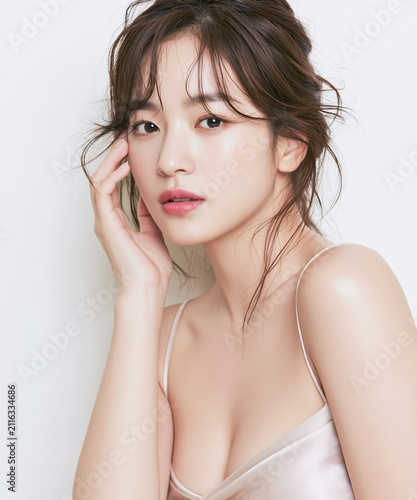 Portrait of young East Asian woman with smooth glowing skin, soft natural makeup, and messy updo hair posing gracefully against bright white studio background