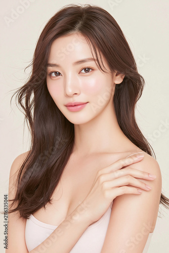 Vertical studio portrait of beautiful young Japanese woman with glowing skin touching shoulder gently looking at camera on soft pastel beige background