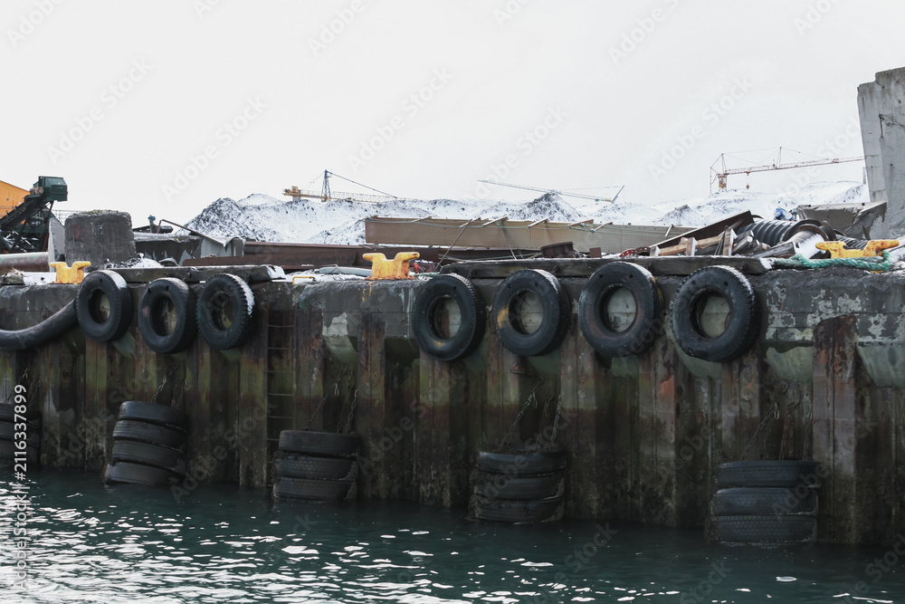 Obraz premium A snowy industrial harbor dock lined with tire fenders and yellow bollards, weathered rusted quay walls and cranes in the background, evoking maritime industry, cold season and urban decay