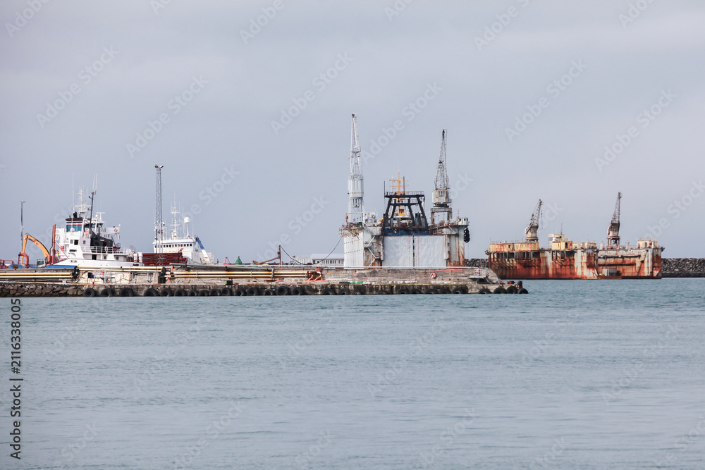 Fototapeta premium Industrial harbor scene with docked ships, rusted cargo vessels and heavy cranes along a cloudy coastal pier, conveying maritime industry, weathered infrastructure and a quiet seafront atmosphere