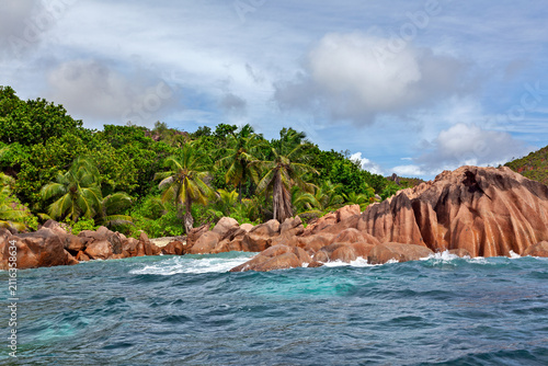 Bild auf Leinwand Tropical shoreline of Curieuse island, Seychelles, where turquoise waves break against sculpted granite rocks beneath coconut palms and lush forest under a cloud-filled sky