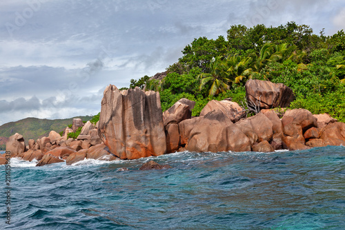 фотографія Tropical shoreline of Curieuse island, Seychelles, where turquoise waves break against sculpted granite rocks beneath coconut palms and lush forest under a cloud-filled sky