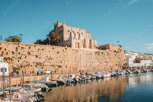 Foto View of the historic Ajuntament de Ciutadella atop harbor walls, with boats moored in calm water and people walking along the promenade in Ciutadella, Balearic Islands, Spain