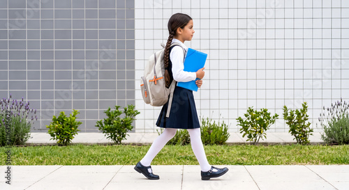 A side view of a young caucasian schoolgirl with a long braid wearing a dark blue uniform and a backpack, walking outside holding a blue folder.