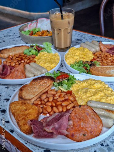 A generous spread of hearty full English breakfast plates with scrambled eggs, sausages, baked beans, and toast served alongside iced coffee at a cafe.
