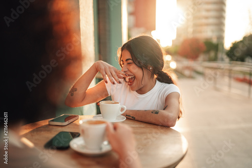 Fotografie Happy young woman laughing, enjoying coffee with friend at street cafe in warm s