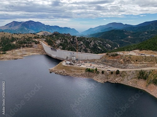 Aerial view of the Gross Reservoir dam construction site featuring the concrete wall, surrounding mountainous terrain, and reservoir water in Boulder County, Colorado, United States.