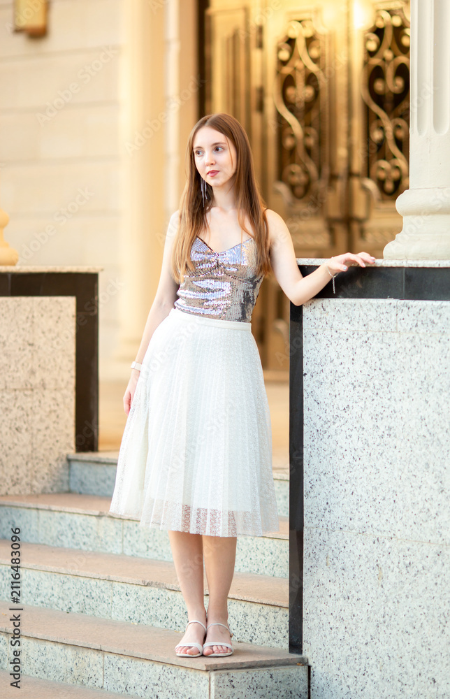 Fototapeta premium An elegant young woman in a stylish outfit poses on the stairs of a classic old building with columns. A fashion portrait, beauty, luxurious lifestyle, confidence, and urban elegance.