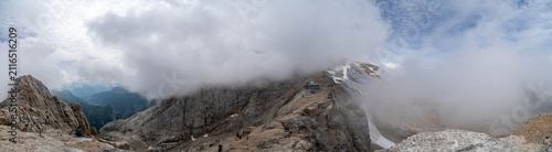 Panoramic aerial view of a lush alpine valley in the Italian Dolomites with towering limestone cliffs, dense forests, winding mountain roads, and dramatic storm clouds under a vibrant blue sky.