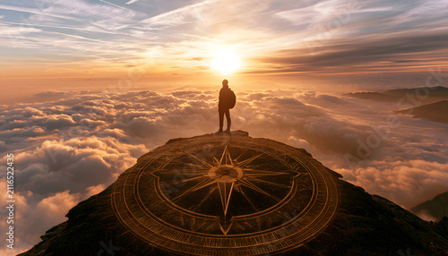 Man silhouette standing on high mountain peak above clouds, looking at golden sunset with a large gold compass glowing on the ground