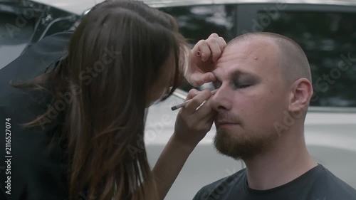 Professional makeup artist applies dark eyeshadow, eyeliner to bearded man during outdoor beauty transformation session for alternative, rock, or theatrical look.
