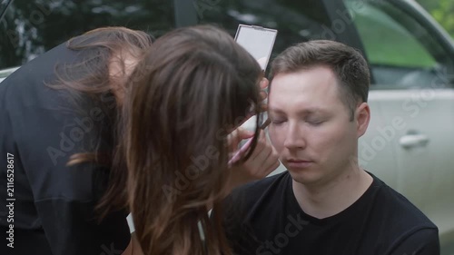 Makeup artist applies dramatic dark makeup to man's face outdoors near vehicle, professional cosmetics application in natural setting.