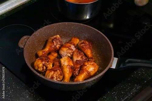 Fotografía Close-up of a hand using a wooden spatula to fry golden brown chicken drumsticks in a dark speckled non-stick pan on a black stovetop