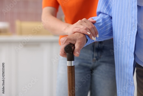 Caregiver supporting elderly woman with walking cane indoors, closeup. Space for text