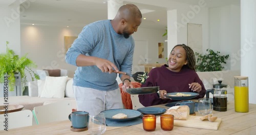 Couple smiling as man leaning, serving eggs with frying pan, spatula at dining table for breakfast