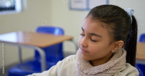 School-age girl glancing at desk, lifting hand to cheek, thinking in classroom wearing knit scarf