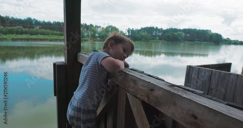 Cute little boy looking down thoughtfully from wooden pier near lake