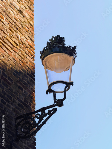 Detailed shot of a vintage black wrought iron wall-mounted street lamp attached to an antique textured brick wall against a clear blue sky.