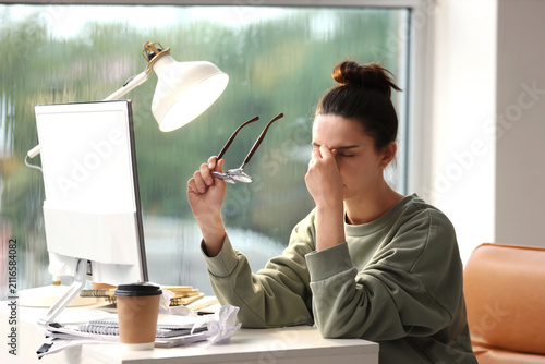 Young woman experiencing eye strain and headache from prolonged computer work at a messy desk, holding her glasses, Challenges of remote work and digital screen time