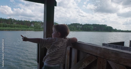 Cute little boy standing on wooden pier and pointing finger towards lake