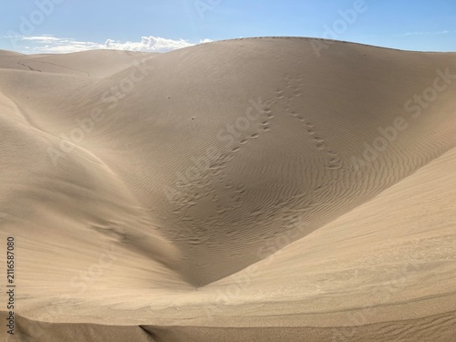 Fotografie Footprints Crossing Wind-Sculpted Sand Dunes Under Blue Sky
